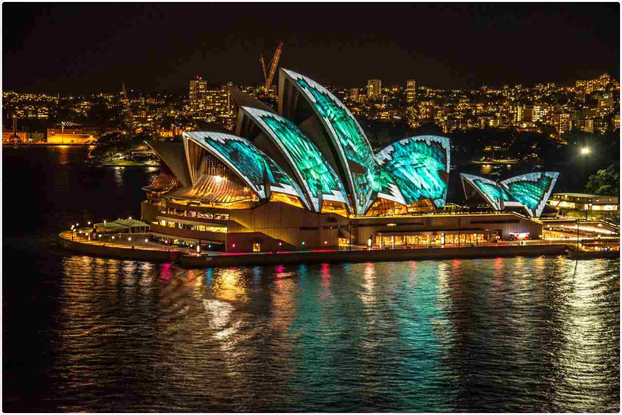 Sydney Opera House illuminated against a dark night sky, reflecting in the harbor waters.